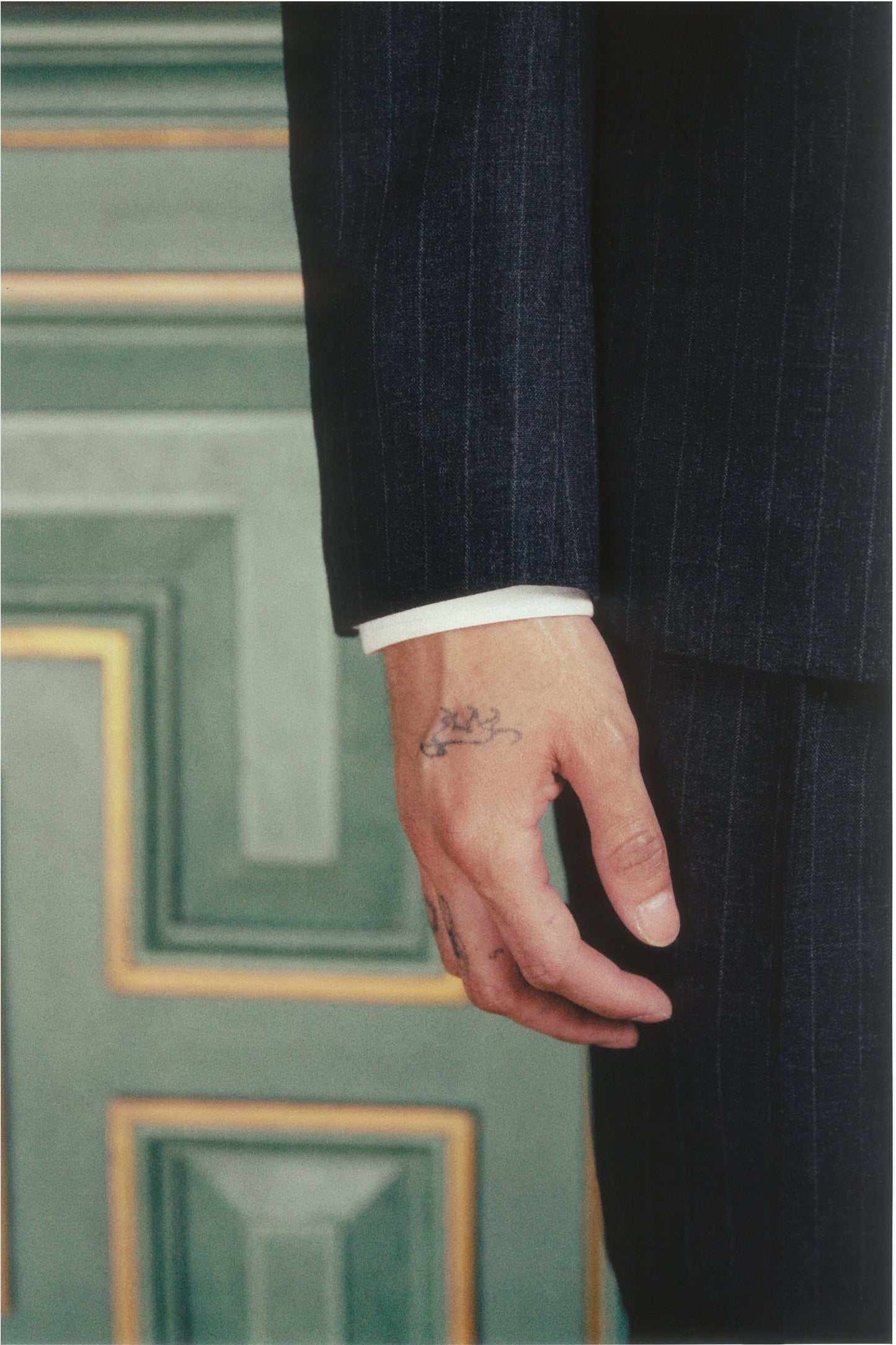 Hand with a tattoo wearing a dark suit sleeve against a green patterned wall.